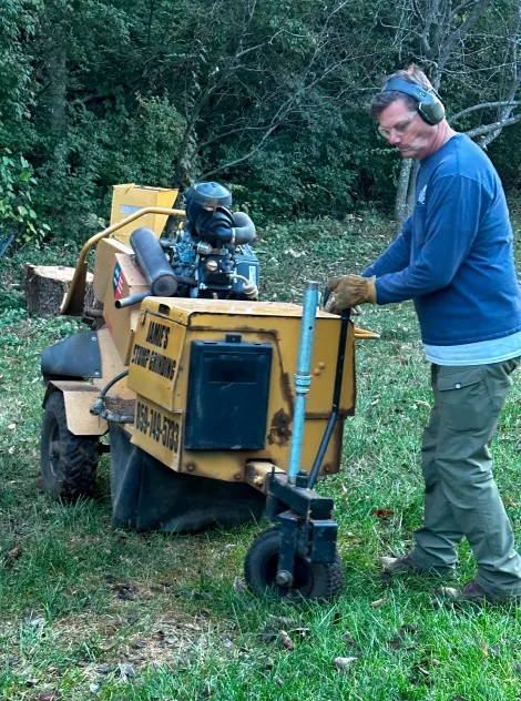 Man wearing ear protection and a blue long sleeve shirt operating a yellow stump grinder outdoors near a treeline, by Jamie's Stump Grinding in Harrodsburg KY